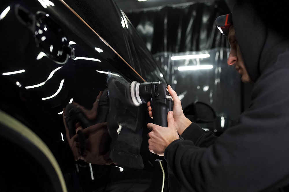 A person in a hoodie uses a power buffer to polish a black car door in a dimly lit garage, showcasing the expert touch of Window Tinting and Detailing Services in New Orleans.