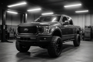 A lifted Ford F-150 pickup truck with large off-road tires, part of the Custom Cars in Kenner lineup, is parked inside a garage under bright overhead lights.